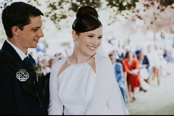 Bride smiles as groom looks at her at the altar, makeup by Jennifer Louise Studio.
